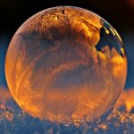 Close-up shot of a frozen bubble with warm reflections resting on a snowy surface at twilight.